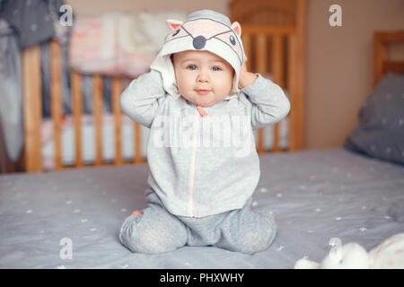 Portrait of cute adorable Caucasian blonde smiling baby girl with blue eyes in grey pajama with fox cat animal hood sitting on bed in bedroom. Happy c Stock Photo