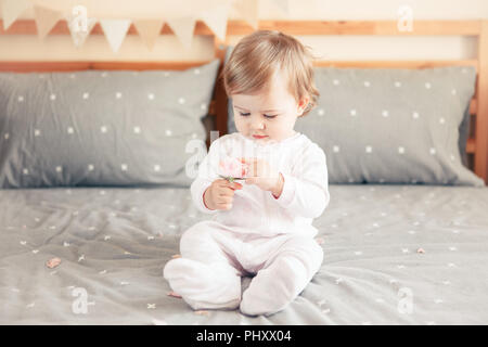 Portrait of cute adorable Caucasian blonde smiling baby girl in white onesie sitting on bed in bedroom and holding pink flower rose. Happy childhood l Stock Photo