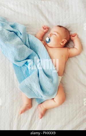 Portrait of a cute adorable white Caucasian baby newborn in diaper, sleeping dreaming with pacifier soother in mouth, lying on bed, covered with blue  Stock Photo