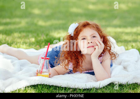 Portrait of cute adorable pensive little red-haired Caucasian girl child in blue dress laying on white blanket in park outside with glass jar of orang Stock Photo