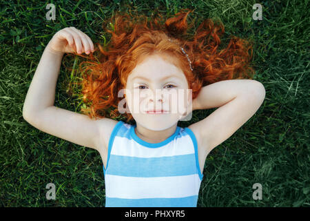 Portrait of cute adorable pensive little red-haired Caucasian girl child in blue dress lying  on green grass in park outside dreaming thinking, happy  Stock Photo