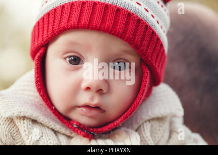 Closeup portrait of cute adorable white Caucasian smiling baby girl boy with large brown eyes in red knitted hat  looking in camera Stock Photo