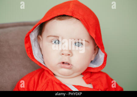 Closeup portrait of cute adorable Caucasian surprised baby boy with blue eyes, wearing red sport hoodie shirt. Seven months child sitting on couch at  Stock Photo