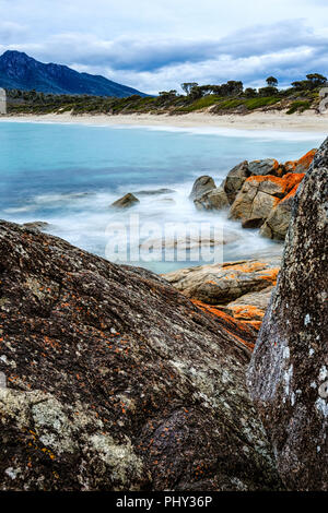 Daytime long exposure capture of Wineglass Bay in Freycinet National Park, Tasmania, Australia, with orange lichen on granite rocks in the foreground. Stock Photo