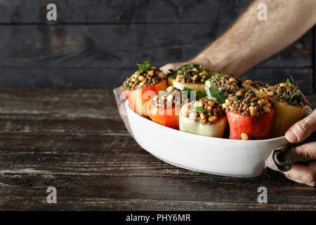 Male hands holding raw green lentils, corn and salsa stuffed peppers in baking dish on wooden table copy space. Healthy, vegetarian food concept Stock Photo