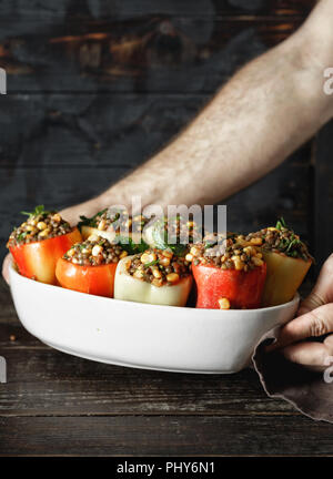 Male hands holding raw green lentils, corn and salsa stuffed peppers in baking dish on wooden table copy space. Healthy, vegetarian food concept Stock Photo