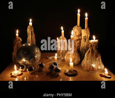 New Year still life : burning candles in glass bottles, crystal candlesticks and sea shells, mirror ball, silver Christmas decorations on a table in the dark. Melted wax, flames, lights, sparkles. Stock Photo