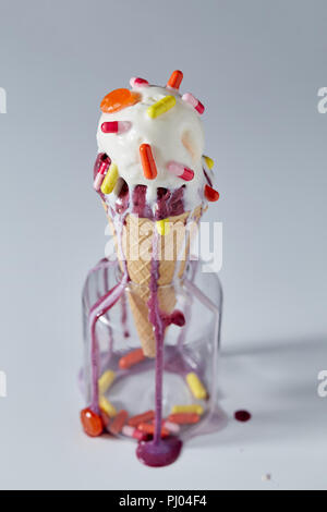 A balls ice cream in a waffle cone with red and yellow tablets and lollipops stands in a glass jar on a gray table. Pils and lollipops in a jar. Stock Photo