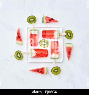 Creative pattern from different pieces of fruit and berry color ice cream on a stick in the shape of a square in a white plate on a gray marble background with a copy space. Flat lay Stock Photo