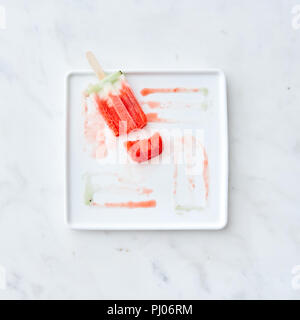 Broken fruit ice cream lolly on a white plate with a pattern from the thawing ice cream on a gray marble background. Copy space for text. Flat lay Stock Photo