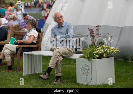 Senior man sitting alone on white bench seat, arms folded & legs crossed, crowd of people beyond - RHS Chatsworth Flower Show, Derbyshire, England, UK Stock Photo