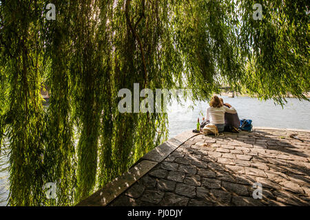 Man and woman sitting close to each other on the edge of the seine river by Square du Vert-Galant in Paris, with a bottle of wine next to them. Stock Photo