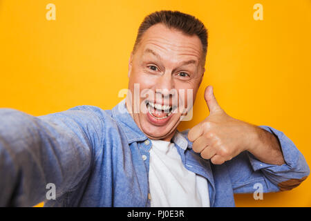Portrait of a confident middle aged man taking a selfie with outstretched hand isolated over yellow background Stock Photo