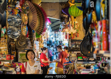 Smiling woman vendor sits in a passage of Ben Thanh Market among shelves with souvenirs: paper hand fans, lanterns, poster of Breaking Bad series, etc Stock Photo