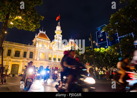 Ho Chi Minh City, Vietnam - May 1, 2018: People drive scooters with headlights on passing by Saigon City Hall, Vincom Center towers and trees at night Stock Photo