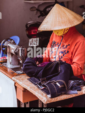 Nha Trang, Vietnam - May 5, 2018: a tailor in a rice hat and a mask (that hide her face) works with jeans at the table with an old sewing machine. Stock Photo