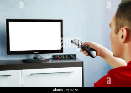 TV remote control in male hand in front of widescreen TV set with blank screen on blue wall background. Young guy switches channels on the remote from Stock Photo