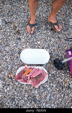 cropped view of male feet in sandals standing in gravel next to barbecue and a plate of raw sausages, frankurters and beef steaks with fresh rosemary Stock Photo