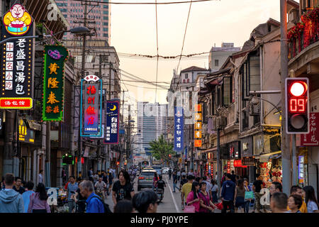 Crowd of local Chinese quickly move along a side street full of colorful signs in Shanghai, China, as the sun sets on a summer day. Stock Photo