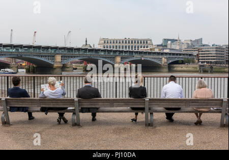 a row of different people male and female sitting on benches looking over the river thames during their lunch break in central London. Stock Photo