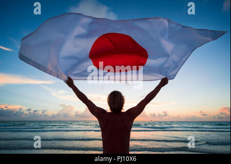 Man holding a fluttering iconic Japanese flag with circle of stars on beach at sunrise Stock Photo