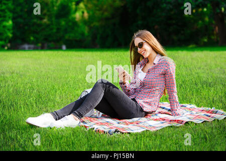 Young pretty woman in plaid shirt wearing sunglasses sitting on the green meadow and writting message on her smartphone Stock Photo