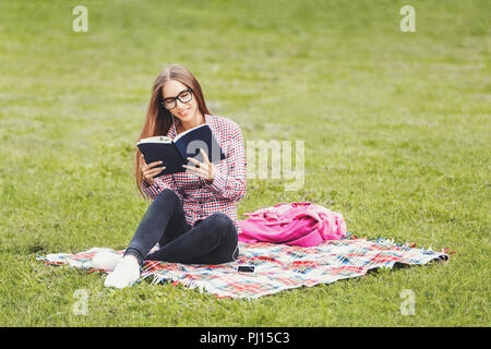 Cute young woman-student in plaid shirt wearing glasses sitting on the green grass and reading her favorite book on the background of green meadow Stock Photo