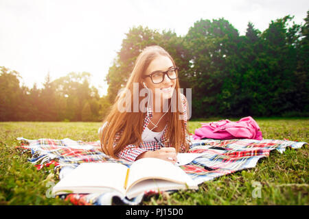 Young girl student in glasses laying on grass with books and studying homework. In the break between lessons she drinks listens to an electronic book Stock Photo