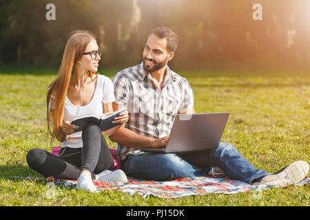 Pretty student-girl in white t-shirt wearing glasses and multicultural student-boy in plaid shirt sitting on the grass, doing homework and speaking with each other Stock Photo