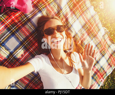 Closeup top view of laughing brunette woman in sunglasses lying on plaid carpet on the grass in park and making selfie Stock Photo