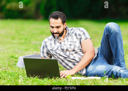 A nice looking black hair man laying on the grass and looking on his laptop. He is doing his homework and preparing for exams Stock Photo