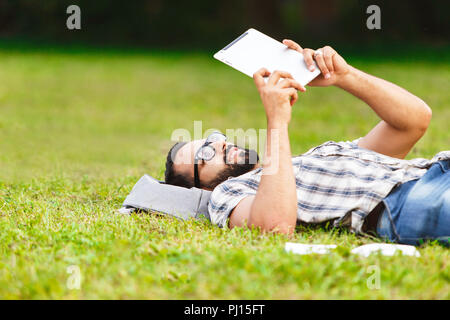 Attractive black hair bearded man in plaid shirt wearing glasses laying on the gray backpack on the grass and reading his favorite book Stock Photo