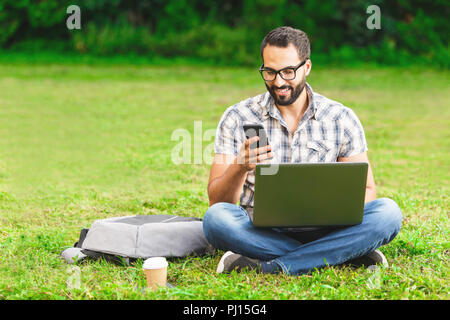 Young bearded man in plaid shirt wearing glasses sitting on the grass in the park with his laptop and writing message Stock Photo