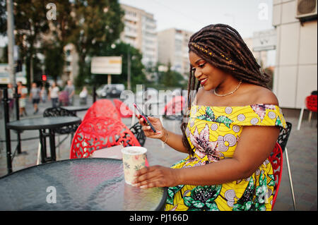 Cute small height african american girl with dreadlocks, wear at coloured yellow dress, sitting at outdoor cafe on red chair and drinking coffee. Stock Photo
