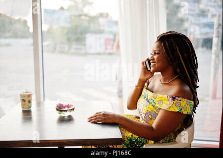 Cute small height african american girl with dreadlocks, wear at coloured yellow dress, sitting at cafe and speaking on phone. Stock Photo