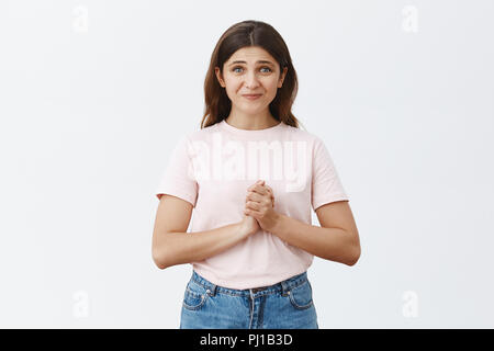 Indoor shot of woman being in need of help clenching hands over chest pursing lips and smiling with intense impatient expression while asking for advice or offer, standing begging over gray background Stock Photo