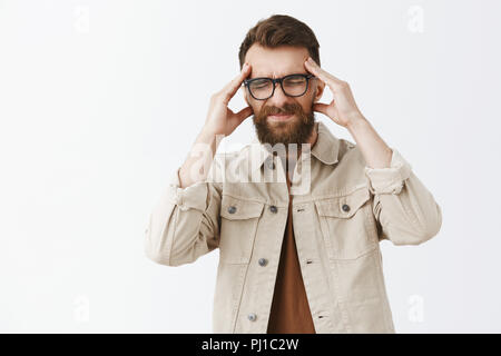 Man losing concentration feeling intense being workaholic grimacing from painful feeling in head holding hands on temples closing eyes and smirking suffering from migraine or headache over gray wall Stock Photo