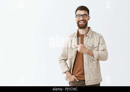 Studio shot of friendly joyful handsome guy with long beard and stylish moustache in trendy glasses and outdoor beige jacket laughing out loud having funny and interesting conversation pointing left Stock Photo