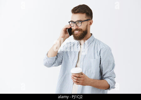 Stylish creative businessman with long beard and cool moustache talking on smartphone turning left smiling with delighted smile having great deal with business partner drinking coffee from paper cup Stock Photo