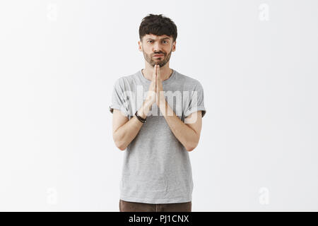 Portrait of questioned doubtful attractive young european man with stylish beard and hairstyle frowning and smirking holding hands in pray while making up mind or thinking about work project Stock Photo
