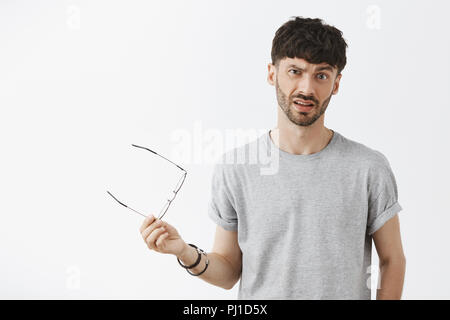 What the heck. Portrait of displeased confused young dark-haired businessman taking off glasses and waving eyewear in hand, frowning from displeased and intense feelings over gray wall Stock Photo