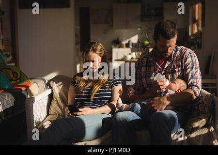 Mother using mobile phone while father feeding his baby on sofa Stock Photo