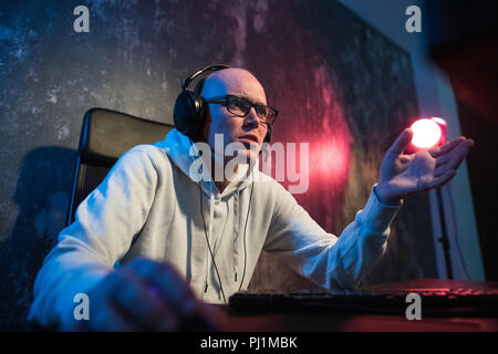 Young man in red headphones playing game at home and streaming playthrough or walkthrough video. Crazy guy is shouting because of unjustice in virtual game and pointing at the screen with one finger. Stock Photo