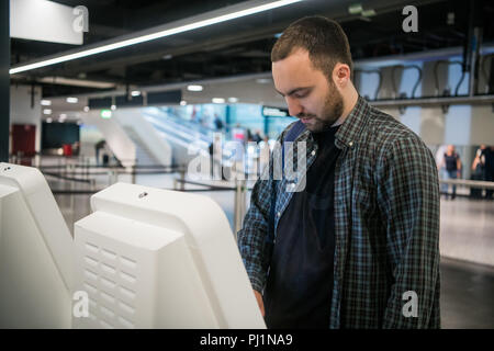 Young man with backpack touching interactive display using self service machine, doing self-check-in for flight or buying airplane tickets at automatic device in modern airport terminal building Stock Photo