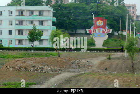 A man cycling past workers homes and a propaganda encouragement billboard in a large town by the rice fields of North Korea Stock Photo