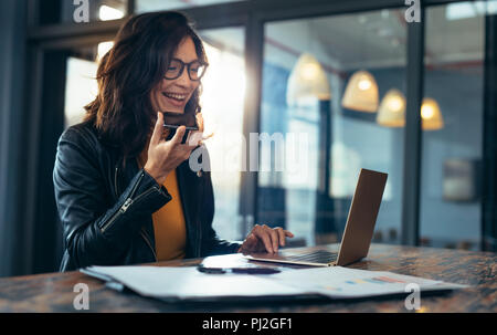 Happy business woman talking on cell phone sitting at table and working on laptop. Asian female sitting at office having telephonic conversation with  Stock Photo