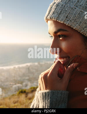 Close up of smiling african woman enjoying the beautiful sea view from mountain top. Female in warm jacket and knitted cap. Stock Photo