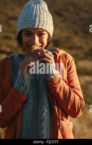 Portrait of cute young woman in winter wear smelling a flower. Female in warm clothes standing on mountain and smiling with a flower in hand. Stock Photo