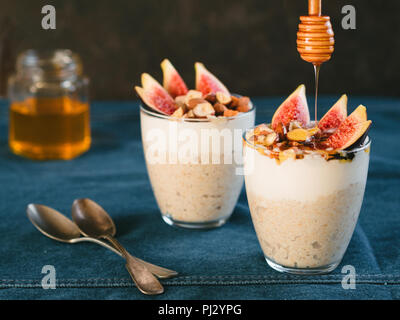 Overnight oats with yogurt,figs and nuts in glass on jeans tablecloth and black background. Idea recipes for healthy breakfast,snack,dessert. Honey drips into glass with overnight oatmeal. Copy space Stock Photo