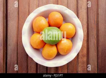 A round white bowl of six mandarin oranges and one green lime sits on a wood slat background. Stock Photo